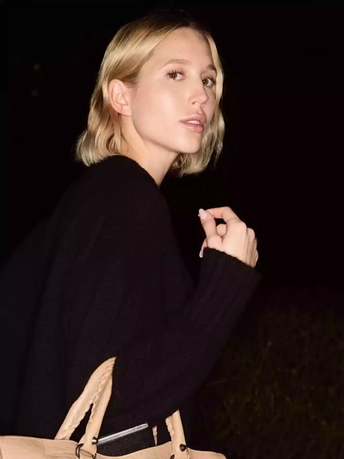 Una persona con cabello rubio corto y suéter negro sostiene una bolsa beige mientras mira hacia atrás en un entorno oscuro. Una persona con cabello rubio corto y suéter negro sostiene una bolsa beige mientras mira hacia atrás en un entorno oscuro.