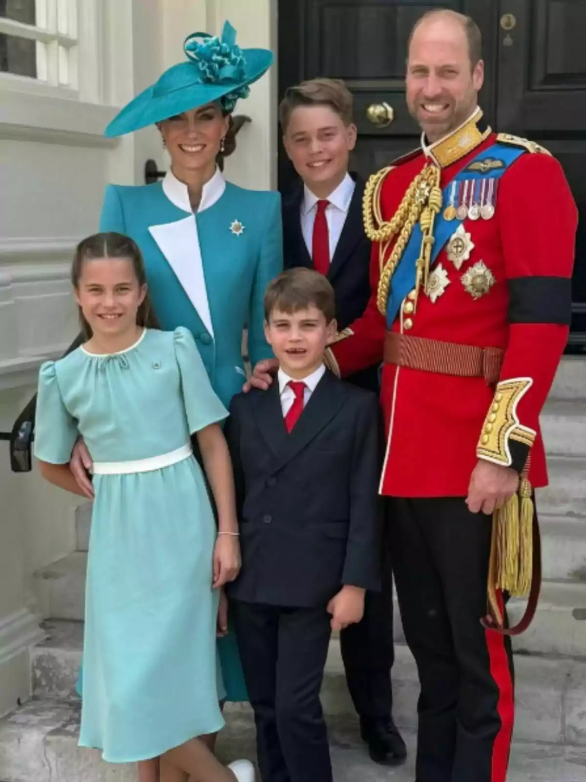 Una família elegantament vestida posa somrient a l’entrada d’un edifici, amb l’home portant un uniforme vermell amb medalles i la dona un vestit blau amb barret a joc, acompanyats de tres nens ben arreglats. Una família elegantament vestida posa somrient a l’entrada d’un edifici, amb l’home portant un uniforme vermell amb medalles i la dona un vestit blau amb barret a joc, acompanyats de tres nens ben arreglats.