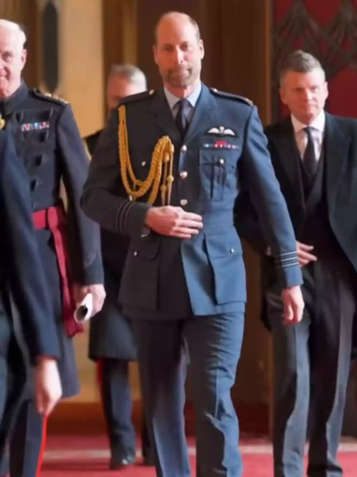 El príncep Guillem amb uniforme militar blau camina per un passadís enmoquetat acompanyat d'altres homes vestits de manera formal. El príncep Guillem amb uniforme militar blau camina per un passadís enmoquetat acompanyat d'altres homes vestits de manera formal.