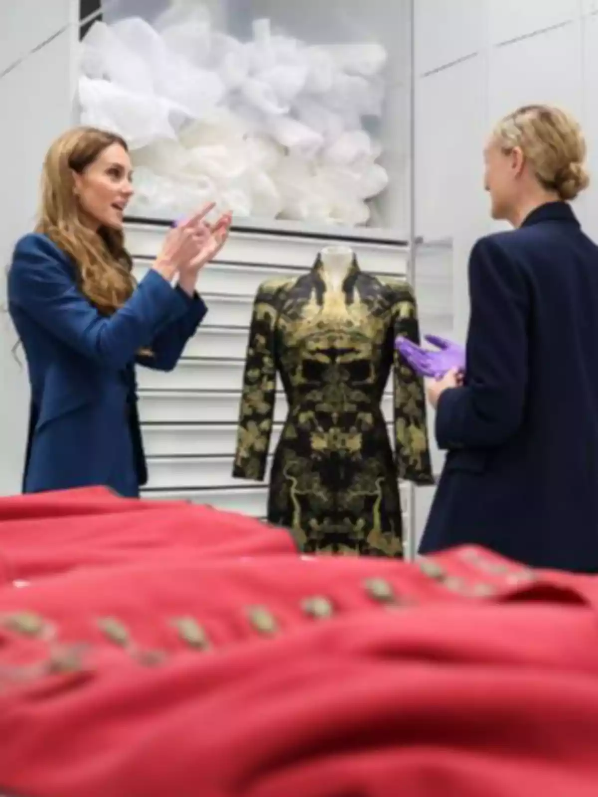 Kate Middleton i una altra dona conversen davant d’un maniquí amb un vestit negre i daurat en una sala on hi ha diverses peces de roba vermelles sobre una taula. Kate Middleton i una altra dona conversen davant d’un maniquí amb un vestit negre i daurat en una sala on hi ha diverses peces de roba vermelles sobre una taula.