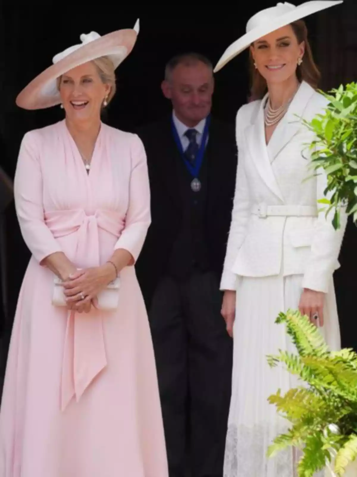 Kate Middleton and another elegantly dressed woman wearing light-colored hats smile outdoors next to a man in a suit in the background. Kate Middleton and another elegantly dressed woman wearing light-colored hats smile outdoors next to a man in a suit in the background.
