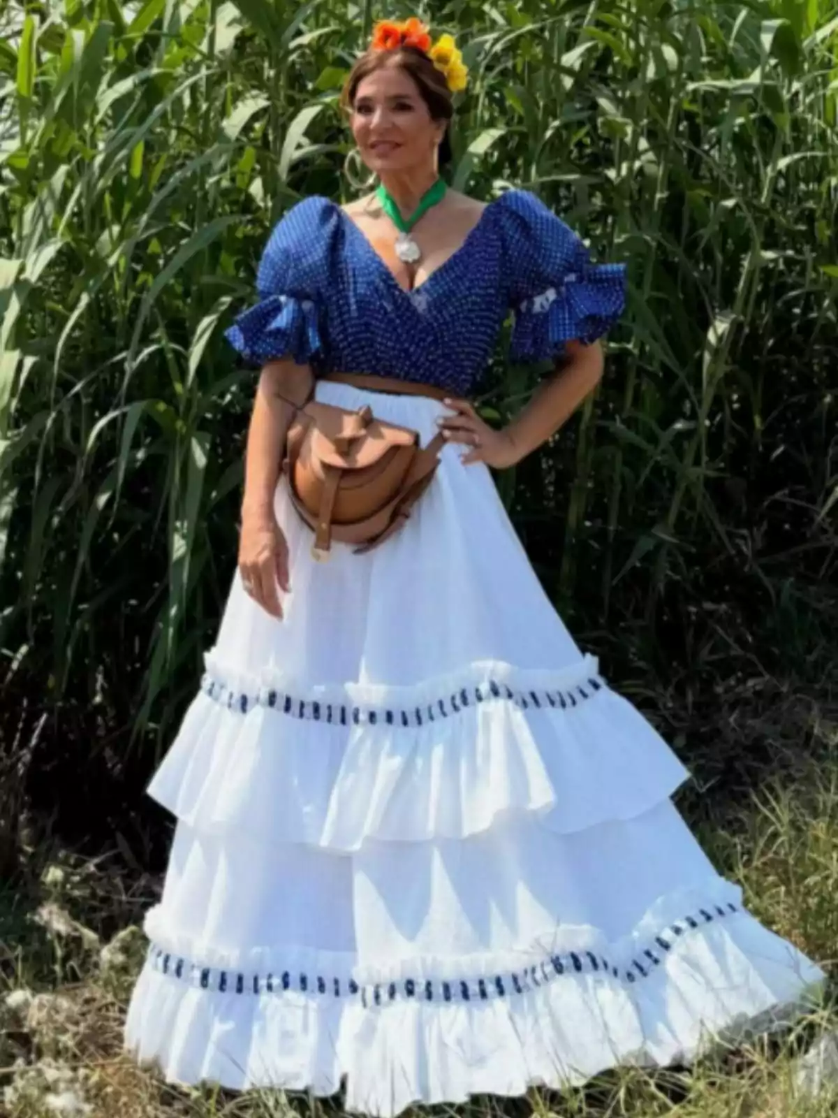 Raquel Bollo vestida amb una faldilla blanca de volants i una brusa blava amb punts, porta flors als cabells i una bossa marró a la cintura, posant a l’aire lliure davant de plantes verdes. Raquel Bollo vestida amb una faldilla blanca de volants i una brusa blava amb punts, porta flors als cabells i una bossa marró a la cintura, posant a l’aire lliure davant de plantes verdes.