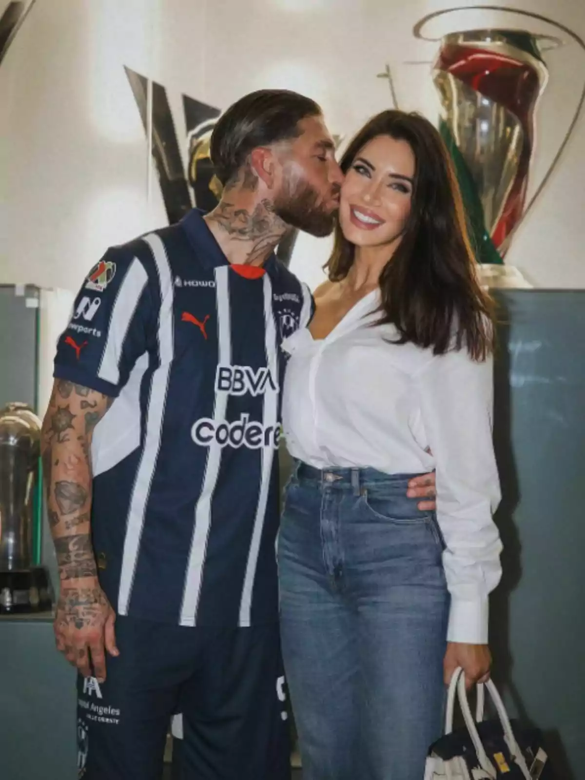 Sergio Ramos amb uniforme de futbol fa un petó a la galta a Pilar Rubio que somriu, tots dos posen davant de trofeus. Sergio Ramos amb uniforme de futbol fa un petó a la galta a Pilar Rubio que somriu, tots dos posen davant de trofeus.