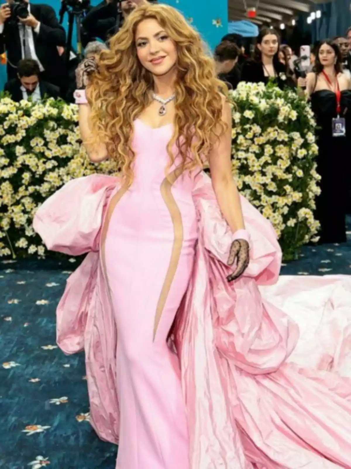 Shakira in a long pink dress poses at an event surrounded by flowers and other people in the background. Shakira in a long pink dress poses at an event surrounded by flowers and other people in the background.