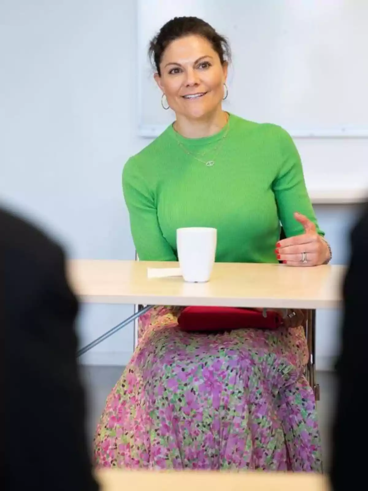 Princess Victoria of Sweden sitting at a table with a white cup, wearing a green sweater and a floral skirt. Princess Victoria of Sweden sitting at a table with a white cup, wearing a green sweater and a floral skirt.