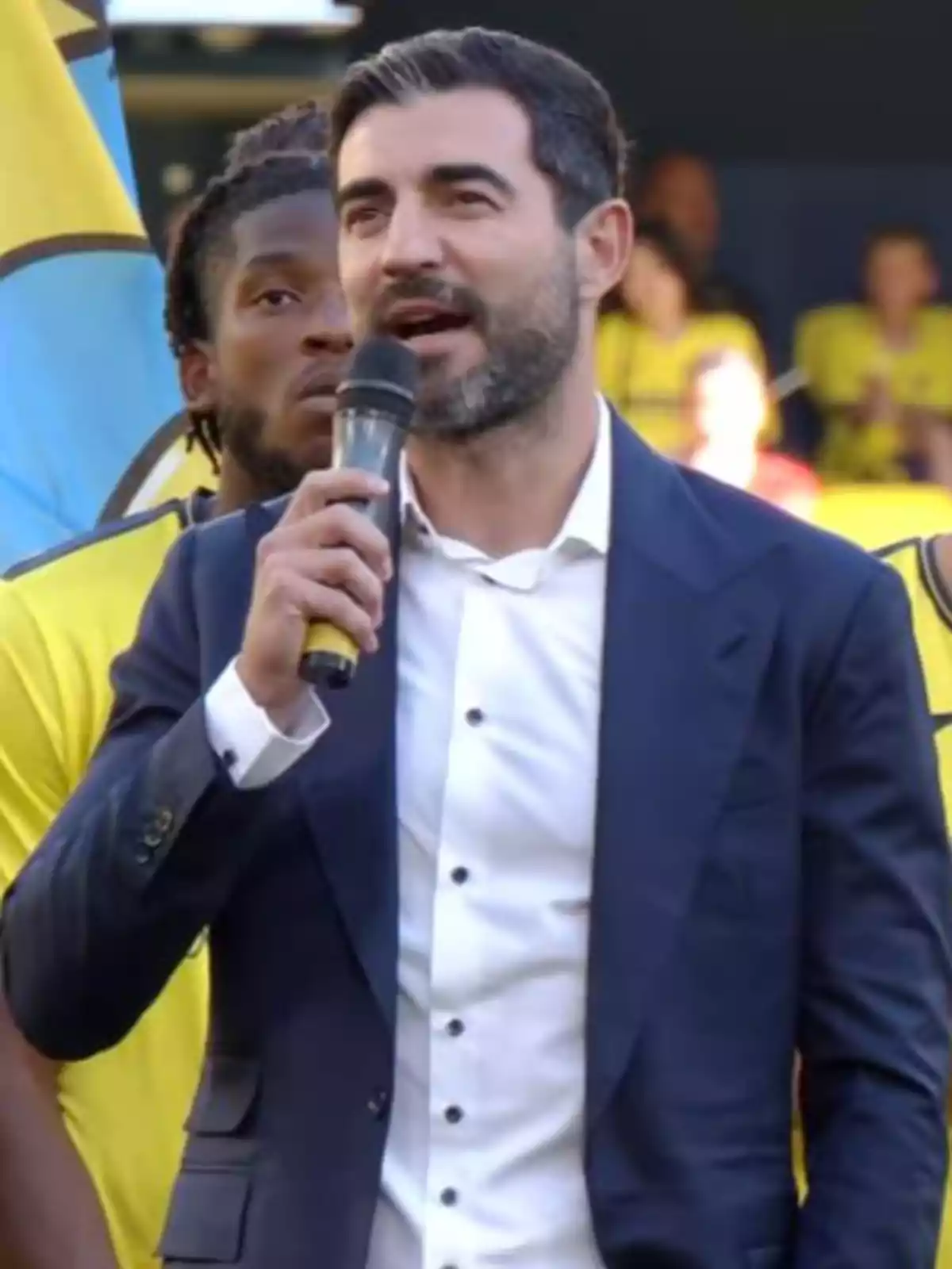 Raúl Albiol in a blue suit speaking into a microphone at a sporting event with people and a yellow flag in the background Raúl Albiol in a blue suit speaking into a microphone at a sporting event with people and a yellow flag in the background