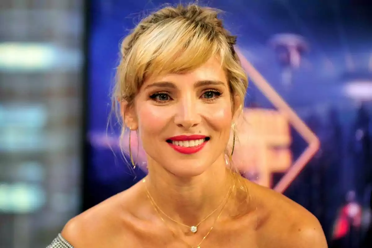 Una persona sonriente con cabello rubio y labios rojos, usando aretes grandes y un collar, con un fondo desenfocado. Una persona sonriente con cabello rubio y labios rojos, usando aretes grandes y un collar, con un fondo desenfocado.