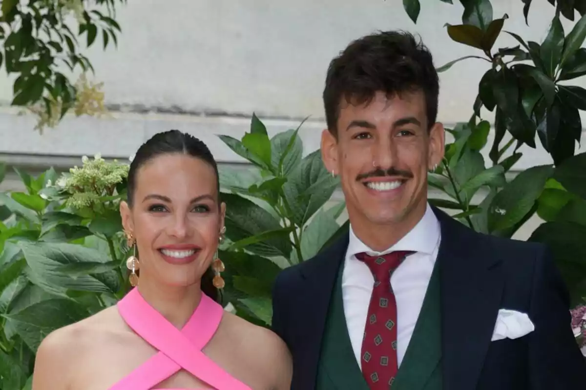 Jessica Bé amb un vestit rosa i Luitingo amb vestit i corbata posen junts davant d'un fons de plantes verdes.