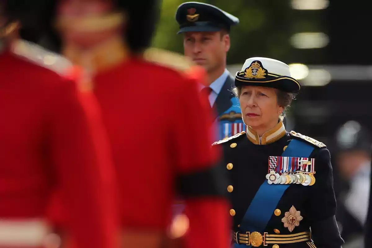 La princesa Ana con uniforme militar y medallas camina junto a el príncipe Guillermo con uniforme de la fuerza aérea, mientras soldados con uniformes rojos están en primer plano. La princesa Ana con uniforme militar y medallas camina junto a el príncipe Guillermo con uniforme de la fuerza aérea, mientras soldados con uniformes rojos están en primer plano.