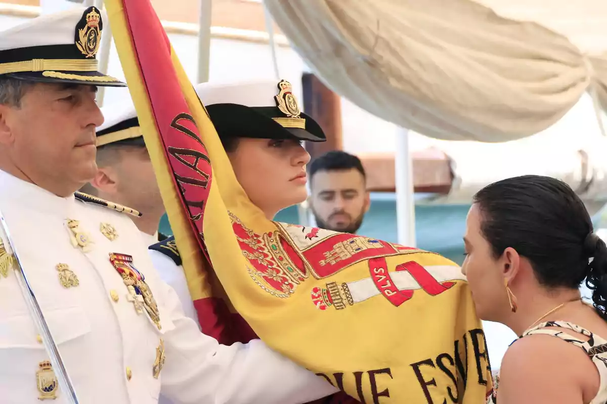 Una cerimònia militar on una persona besa una bandera sostinguda per oficials en uniforme blanc. Una cerimònia militar on una persona besa una bandera sostinguda per oficials en uniforme blanc.