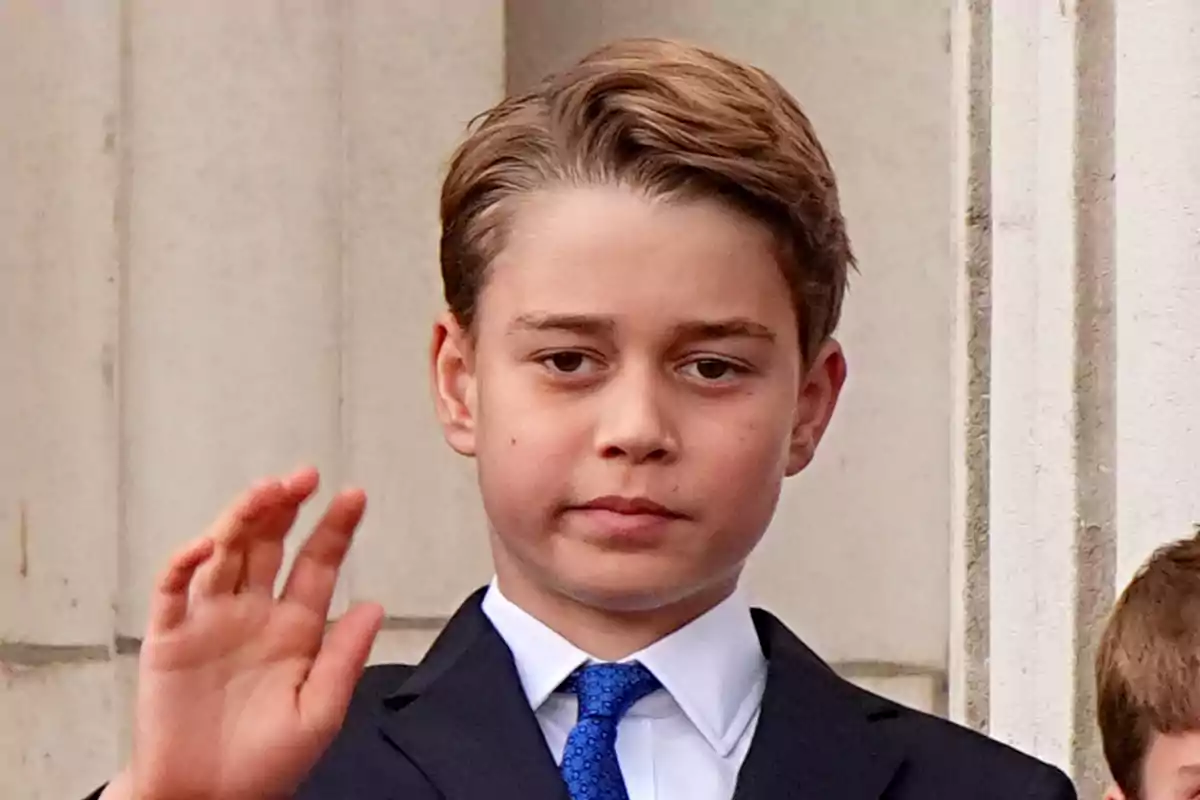 El príncipe George con traje oscuro y corbata azul levanta la mano frente a un edificio de piedra. El príncipe George con traje oscuro y corbata azul levanta la mano frente a un edificio de piedra.
