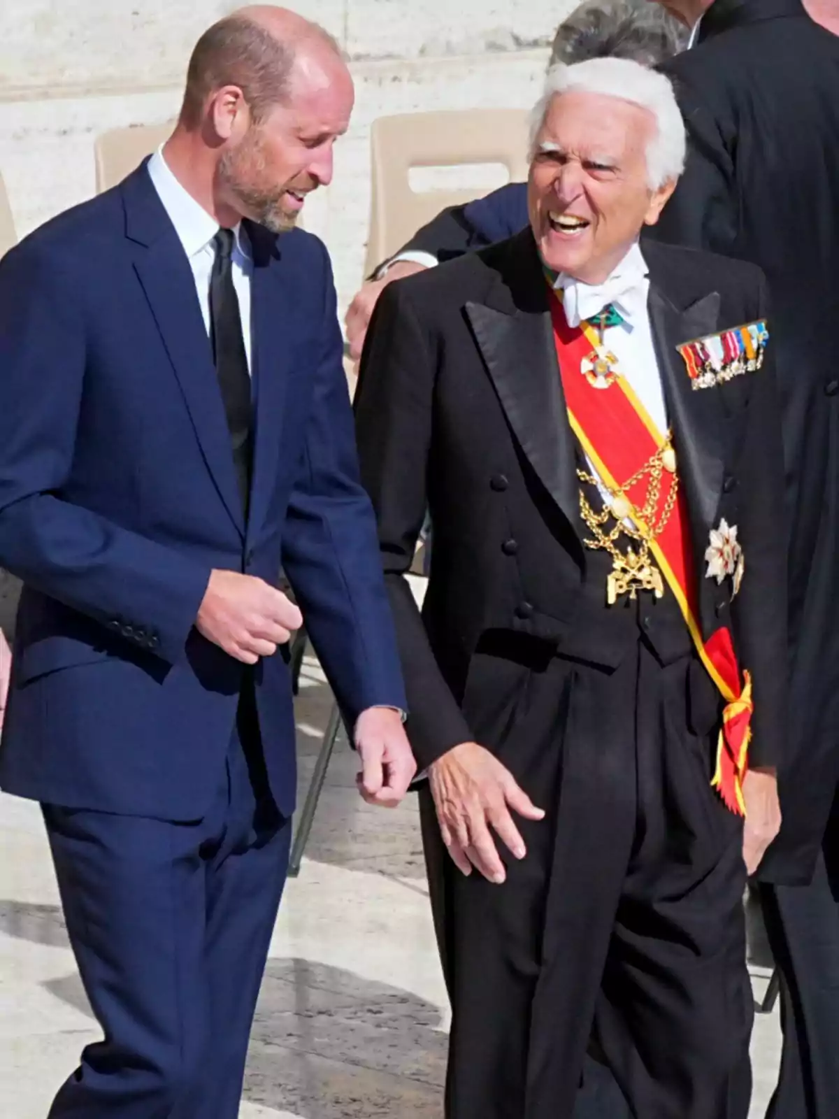 El príncep Guillem i un altre home conversen animadament; un porta un vestit blau i l'altre un vestit formal amb condecoracions i una banda vermella. El príncep Guillem i un altre home conversen animadament; un porta un vestit blau i l'altre un vestit formal amb condecoracions i una banda vermella.