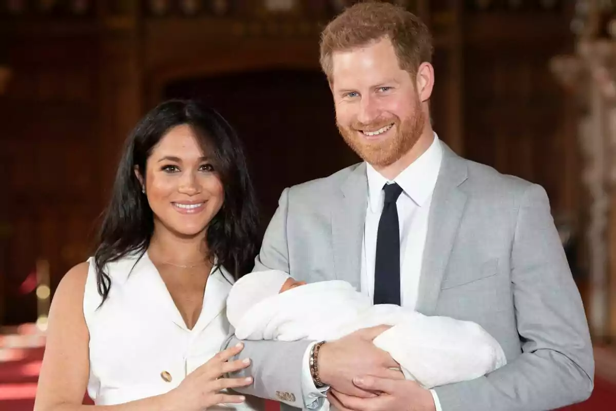 Una parella, formada per Meghan Markle i el príncep Harry, somrient sosté un nadó embolicat en una manta blanca.