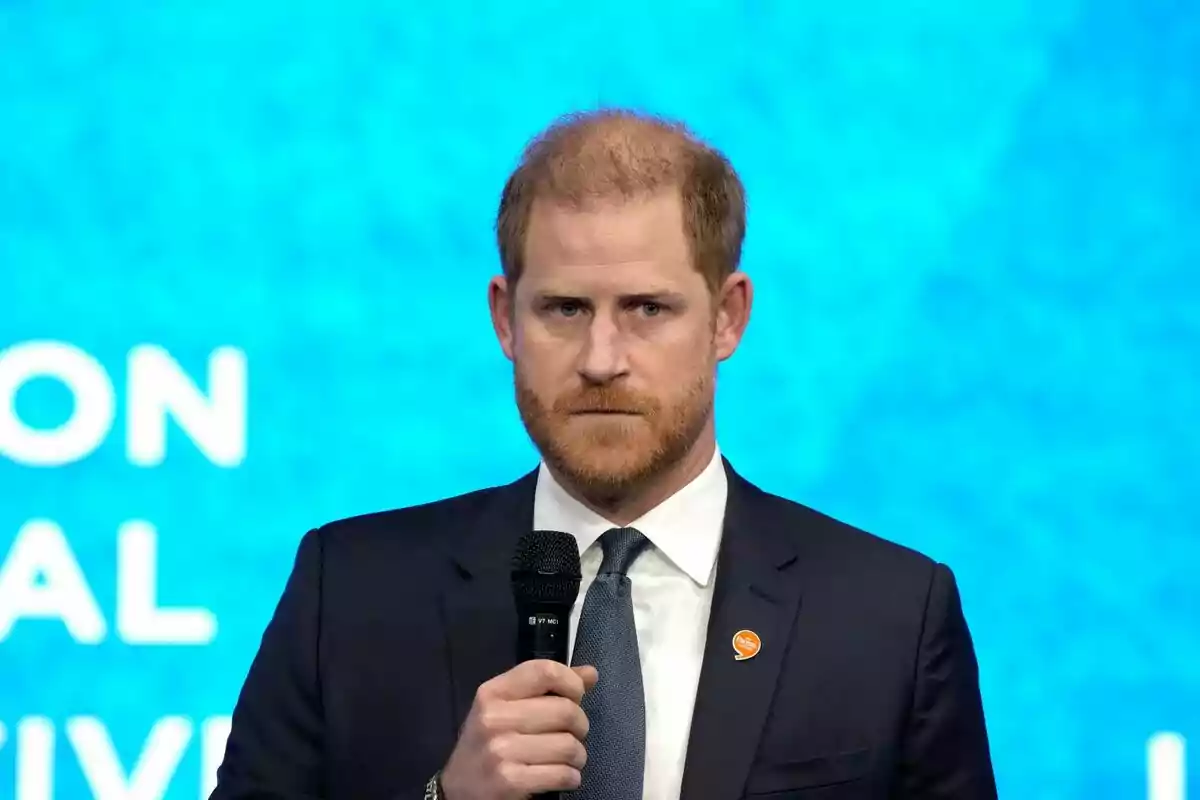 El príncep Harry amb barba i vestit fosc sostenint un micròfon davant d'un fons blau. El príncep Harry amb barba i vestit fosc sostenint un micròfon davant d'un fons blau.