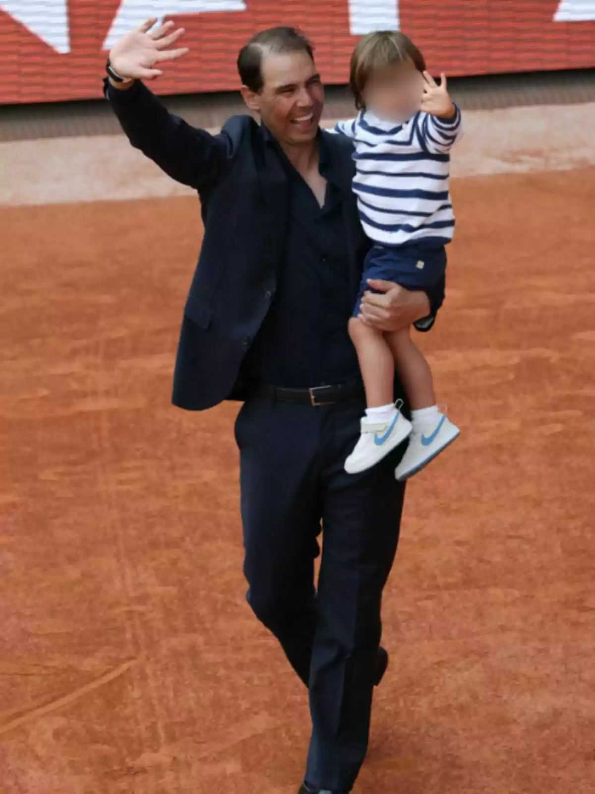 Rafa Nadal sonriente vestido de traje oscuro sostiene a un niño pequeño en brazos mientras ambos saludan con la mano en una cancha de tierra batida Rafa Nadal sonriente vestido de traje oscuro sostiene a un niño pequeño en brazos mientras ambos saludan con la mano en una cancha de tierra batida