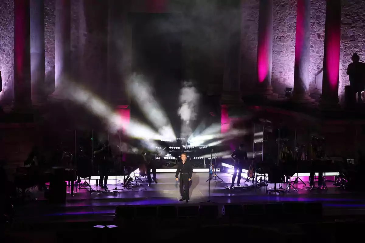 Raphael es presenta en un escenari il·luminat amb llums i fum, acompanyat de músics i envoltat de columnes antigues il·luminades en tons morats. Raphael es presenta en un escenari il·luminat amb llums i fum, acompanyat de músics i envoltat de columnes antigues il·luminades en tons morats.