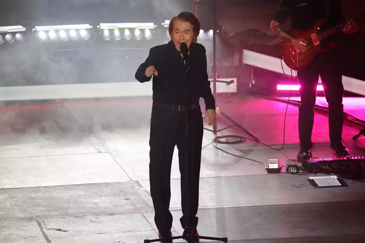 Raphael vestido de negro interpretando una canción en el escenario con luces y humo de fondo mientras un guitarrista lo acompaña Raphael vestido de negro interpretando una canción en el escenario con luces y humo de fondo mientras un guitarrista lo acompaña