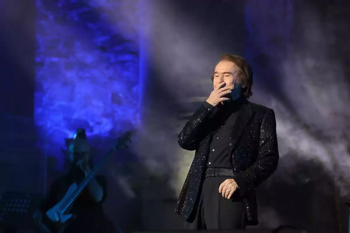 Raphael vestit de negre amb jaqueta brillant damunt l’escenari portant una mà a la boca mentre un músic toca el baix al fons amb llums blaves il·luminant l’escena Raphael vestit de negre amb jaqueta brillant damunt l’escenari portant una mà a la boca mentre un músic toca el baix al fons amb llums blaves il·luminant l’escena