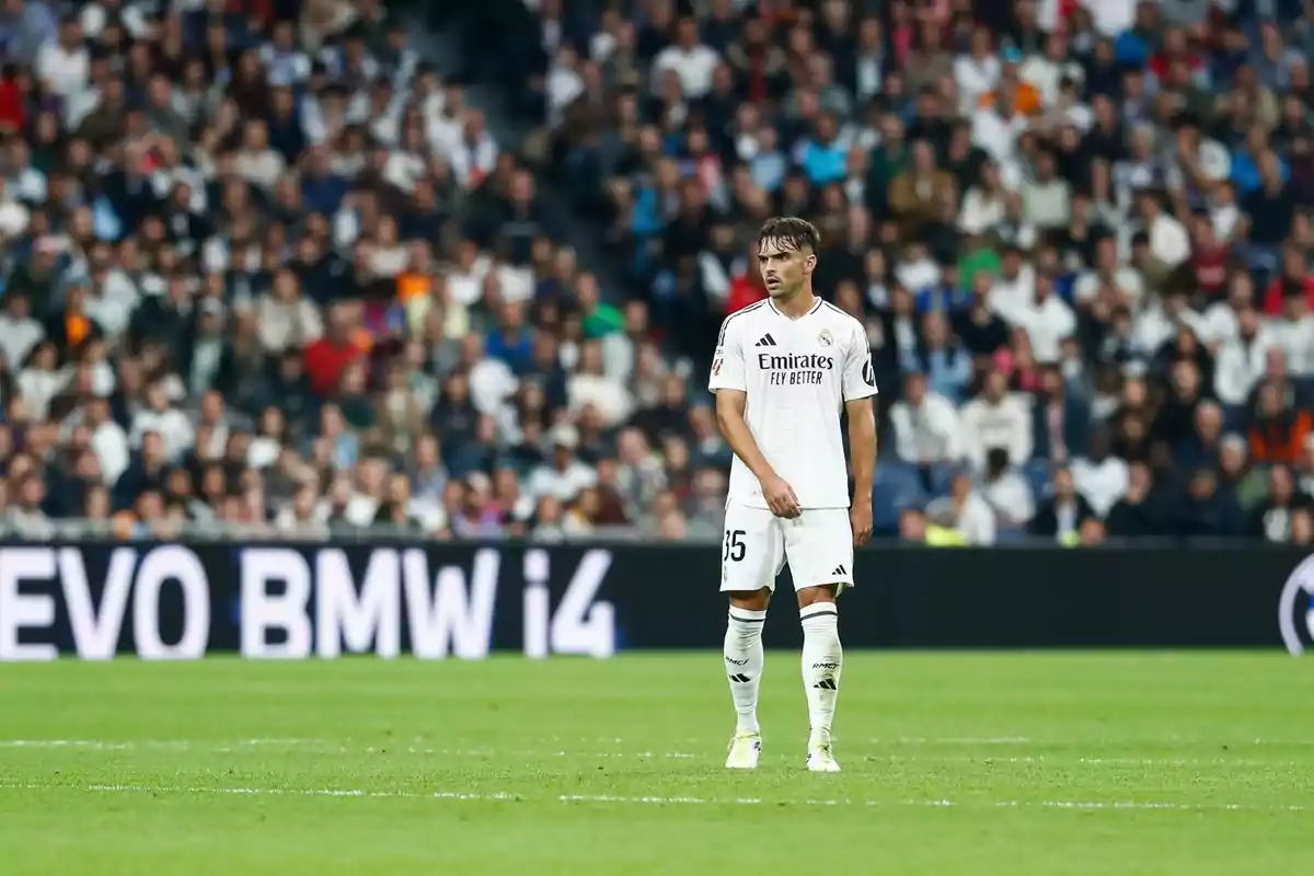 A soccer player in the white uniform of Real Madrid stands on the field during a match, with a crowd of spectators in the background.