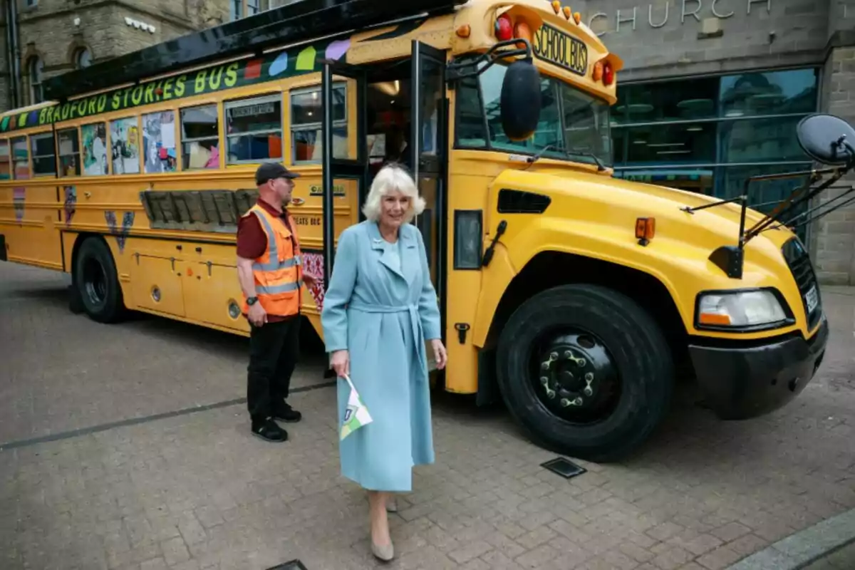 Queen Camila in a light blue coat walks in front of a yellow school bus while a man in an orange vest stands near the bus door. Queen Camila in a light blue coat walks in front of a yellow school bus while a man in an orange vest stands near the bus door.
