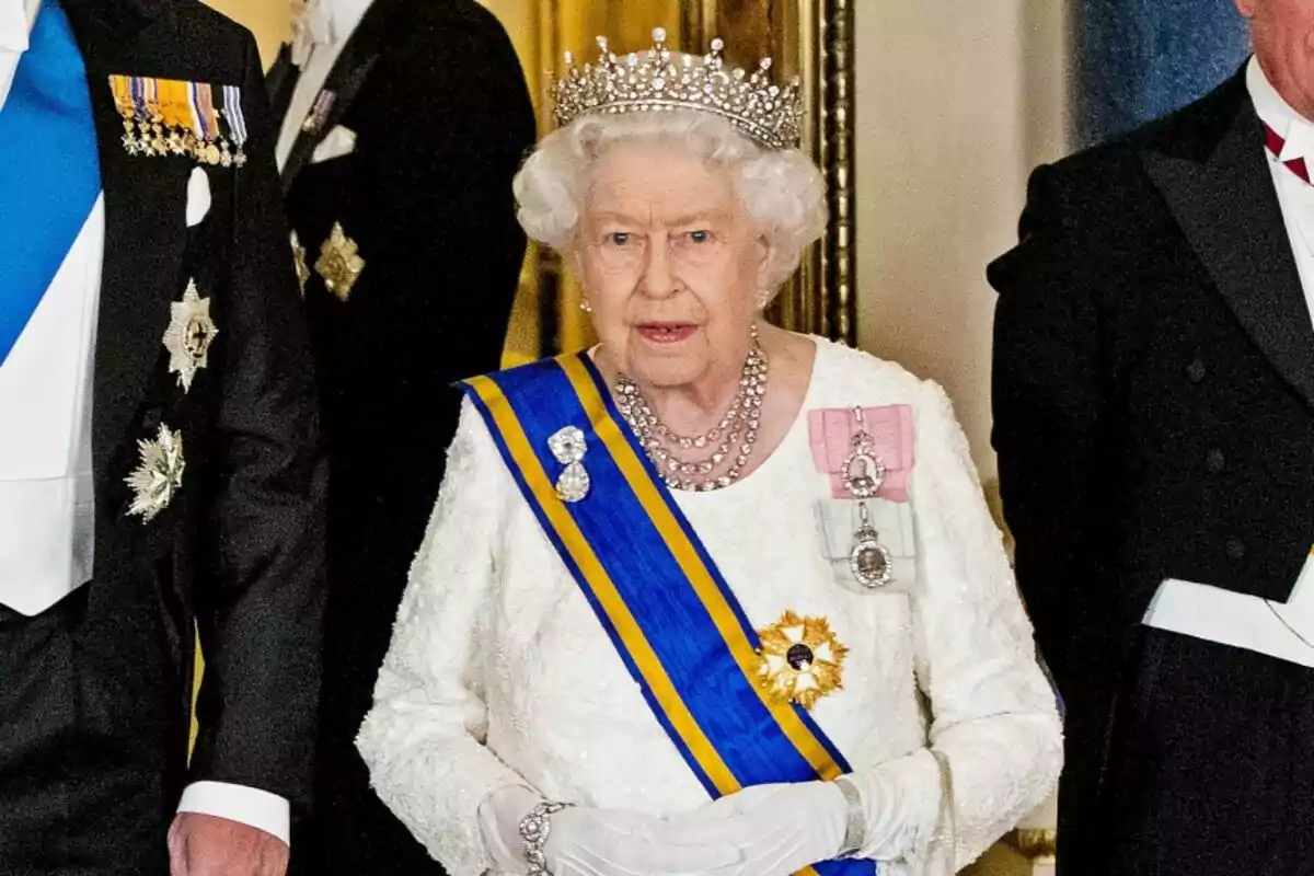 Queen Elizabeth wearing a tiara and formal attire, including a blue and yellow sash, surrounded by people in elegant suits. Queen Elizabeth wearing a tiara and formal attire, including a blue and yellow sash, surrounded by people in elegant suits.
