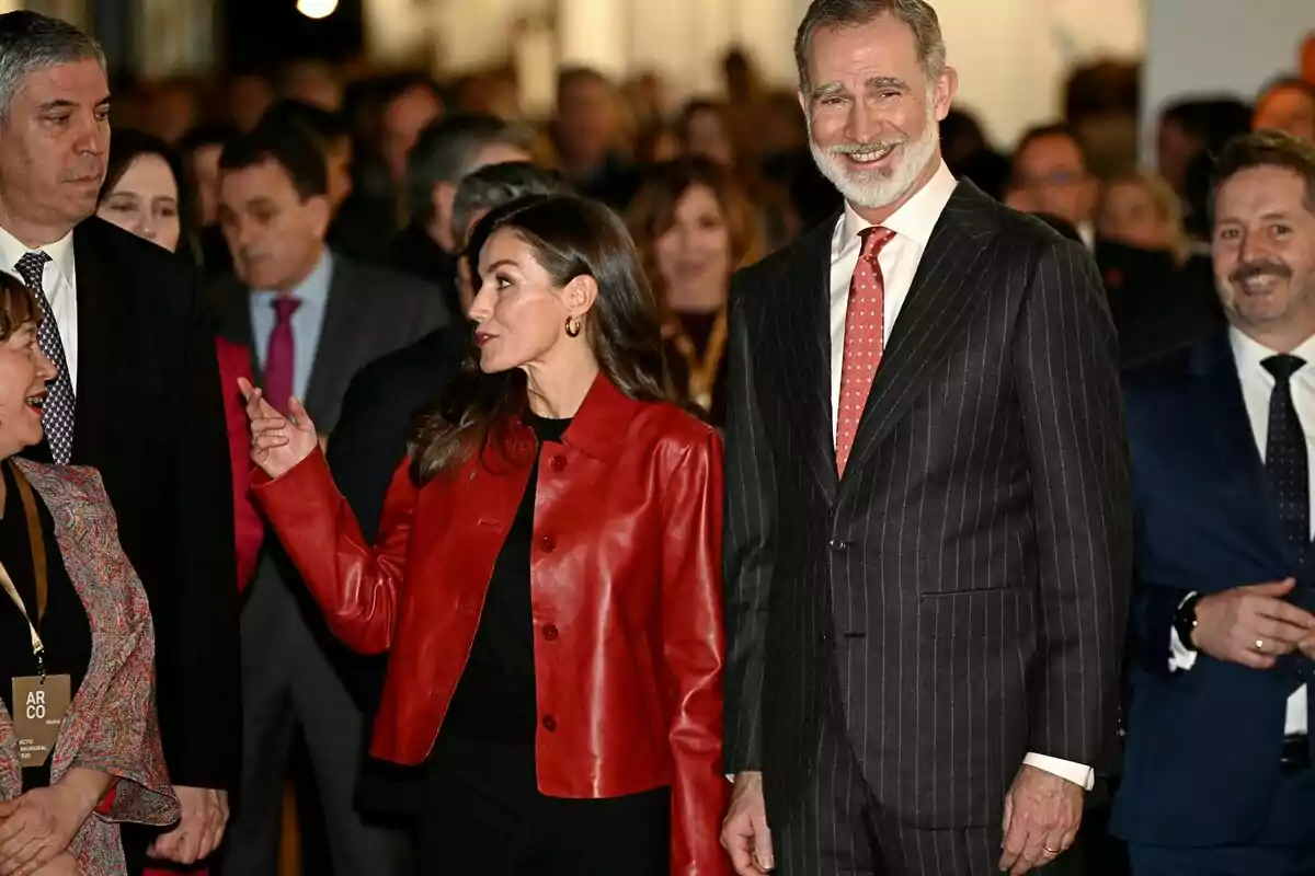 Un grup de persones vestides formalment en un esdeveniment, amb la reina Letícia amb jaqueta vermella parlant i Felip VI amb vestit de ratlles somrient. Un grup de persones vestides formalment en un esdeveniment, amb la reina Letícia amb jaqueta vermella parlant i Felip VI amb vestit de ratlles somrient.