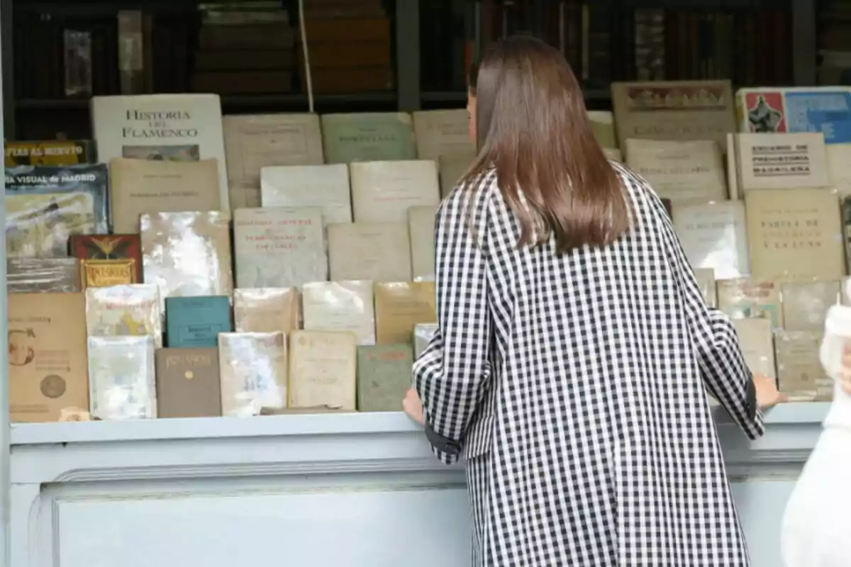 La reina Letizia de espaldas con un abrigo de cuadros observa libros antiguos en una librería al aire libre.