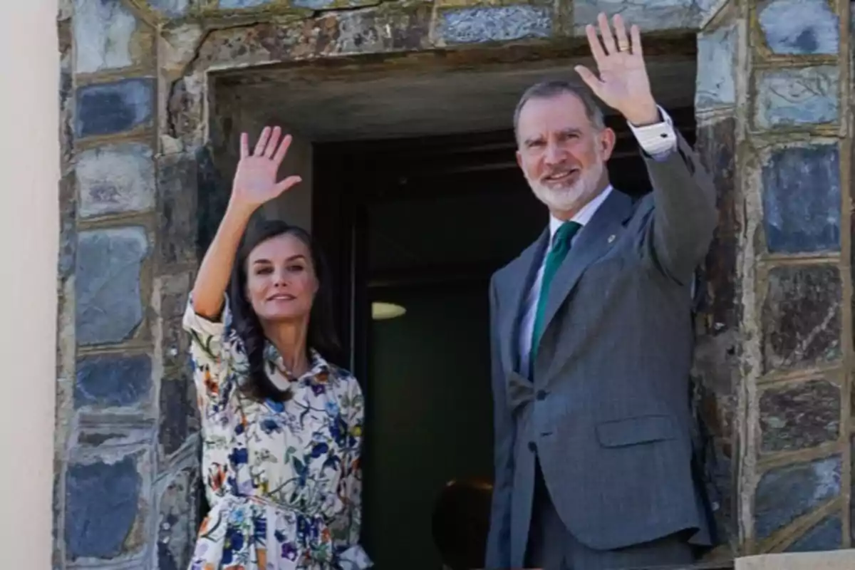 La reina Letizia con vestido floral y Felipe VI con traje gris saludan sonrientes desde una ventana de piedra.