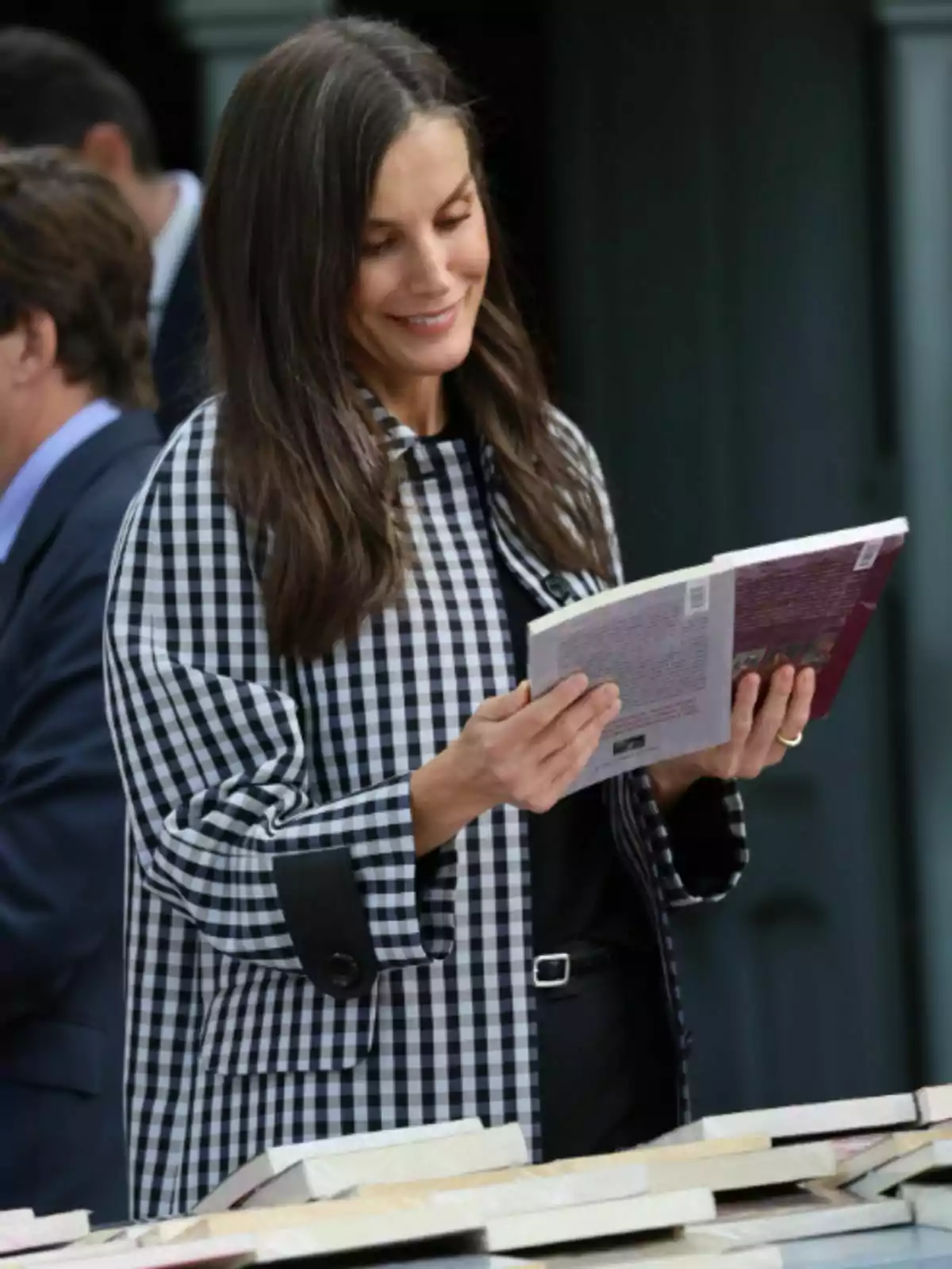 La reina Letizia está leyendo un libro mientras sonríe, vestida con una chaqueta de cuadros blancos y negros, rodeada de más libros sobre una mesa.