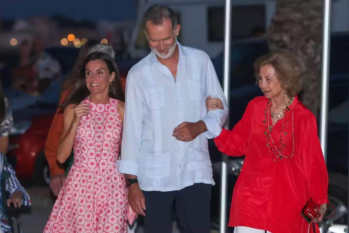 Queen Letizia, Felipe VI, and Queen Sofía smile as they walk together outdoors at night—a woman in a pink dress, a man in a light jersey, and an older woman in a red blouse. Queen Letizia, Felipe VI, and Queen Sofía smile as they walk together outdoors at night—a woman in a pink dress, a man in a light jersey, and an older woman in a red blouse.