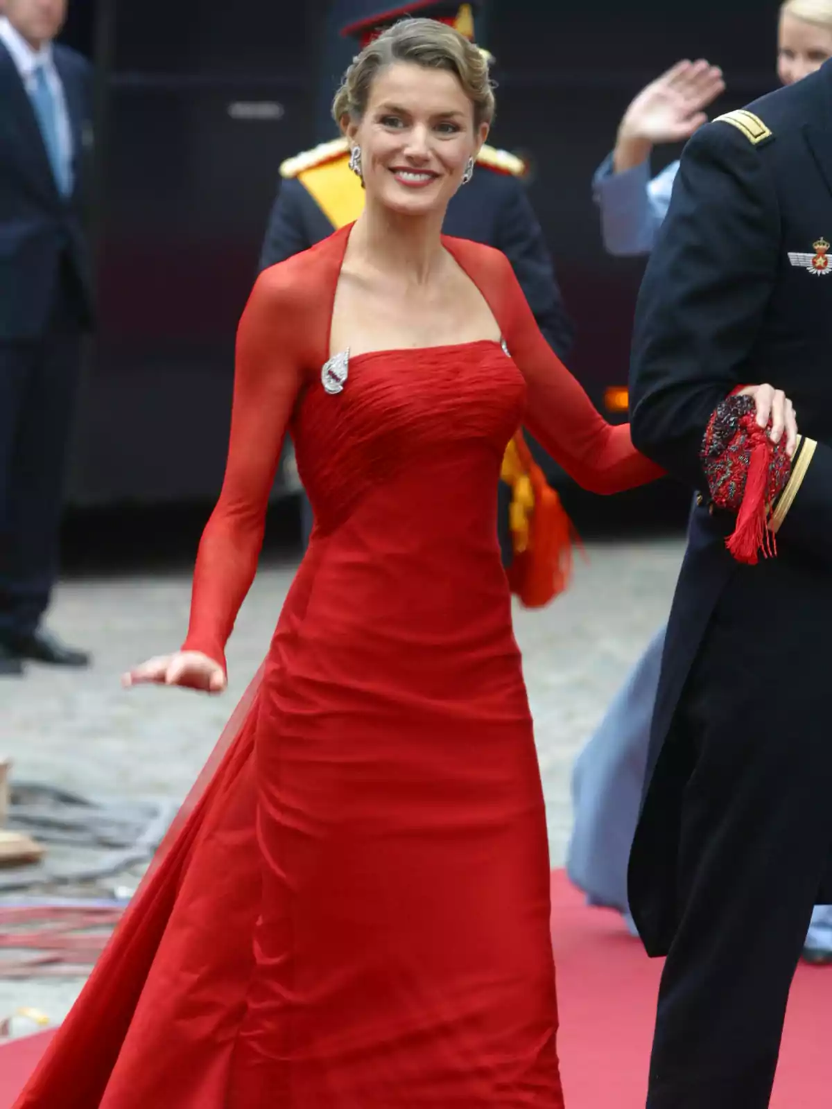 Queen Letizia, smiling in an elegant red gala dress, walks on a red carpet accompanied by a man in military uniform. Queen Letizia, smiling in an elegant red gala dress, walks on a red carpet accompanied by a man in military uniform.