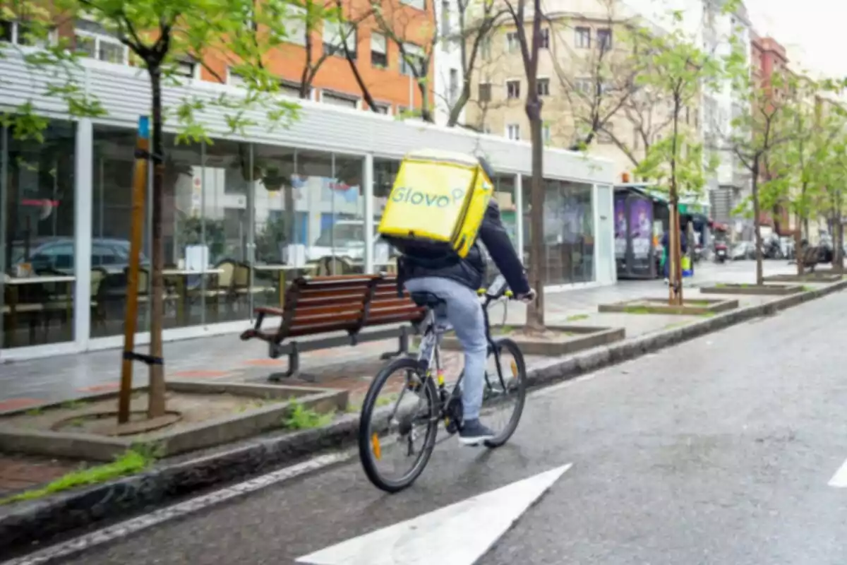 Un repartidor de Glovo amb bicicleta circula per un carrer urbà amb arbres i edificis als costats.