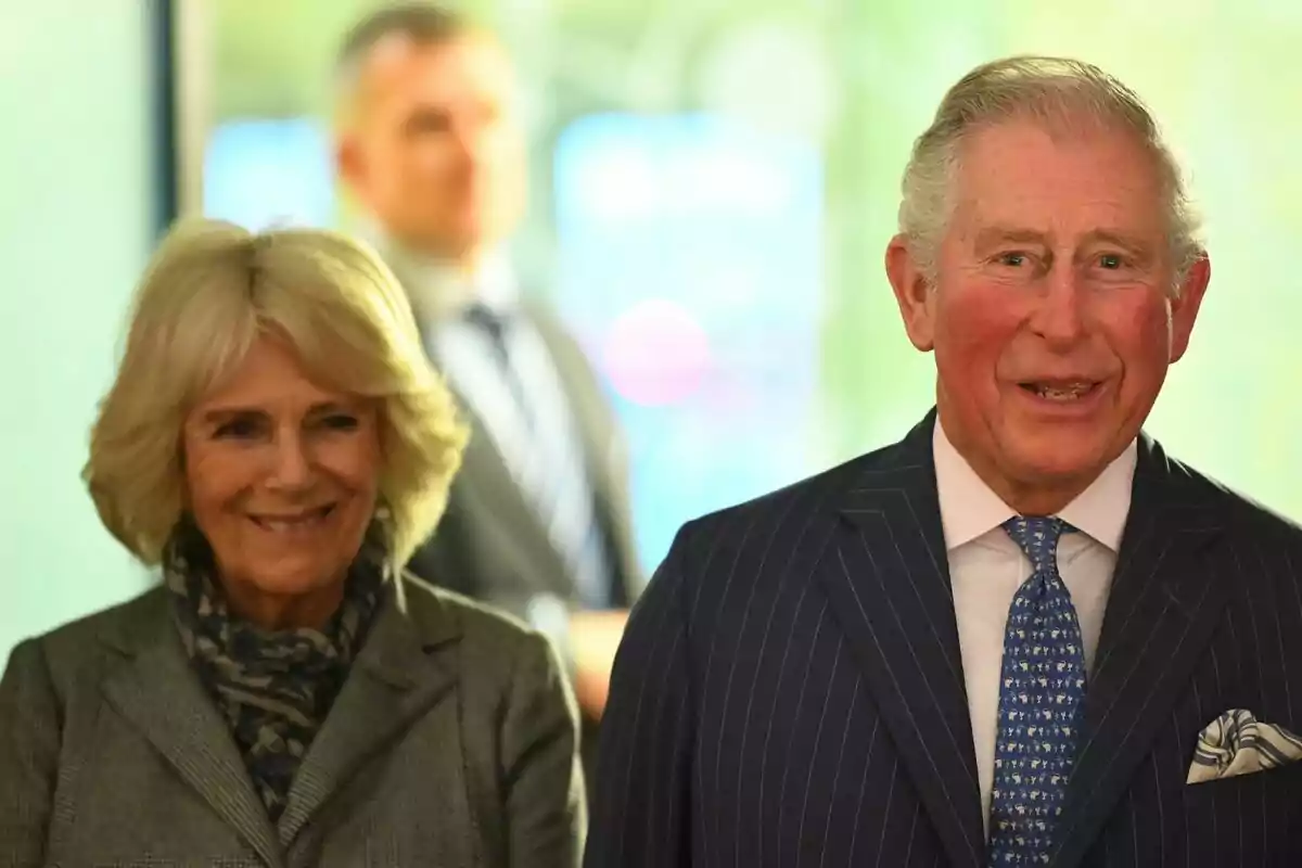 Two elderly people - King Charles III and Camilla Parker - smile while standing, with a blurred background and a third person in the background. Two elderly people - King Charles III and Camilla Parker - smile while standing, with a blurred background and a third person in the background.