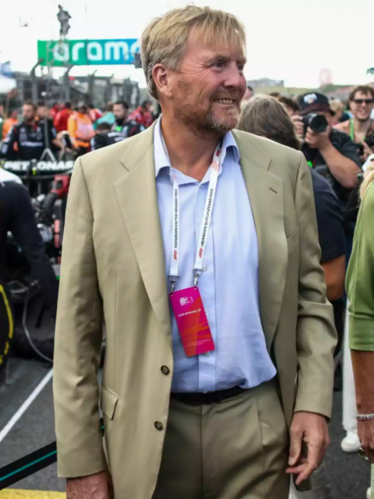 King Willem of the Netherlands in a beige suit and light blue jersey wears a badge around his neck while smiling at an outdoor event surrounded by people and photographers.