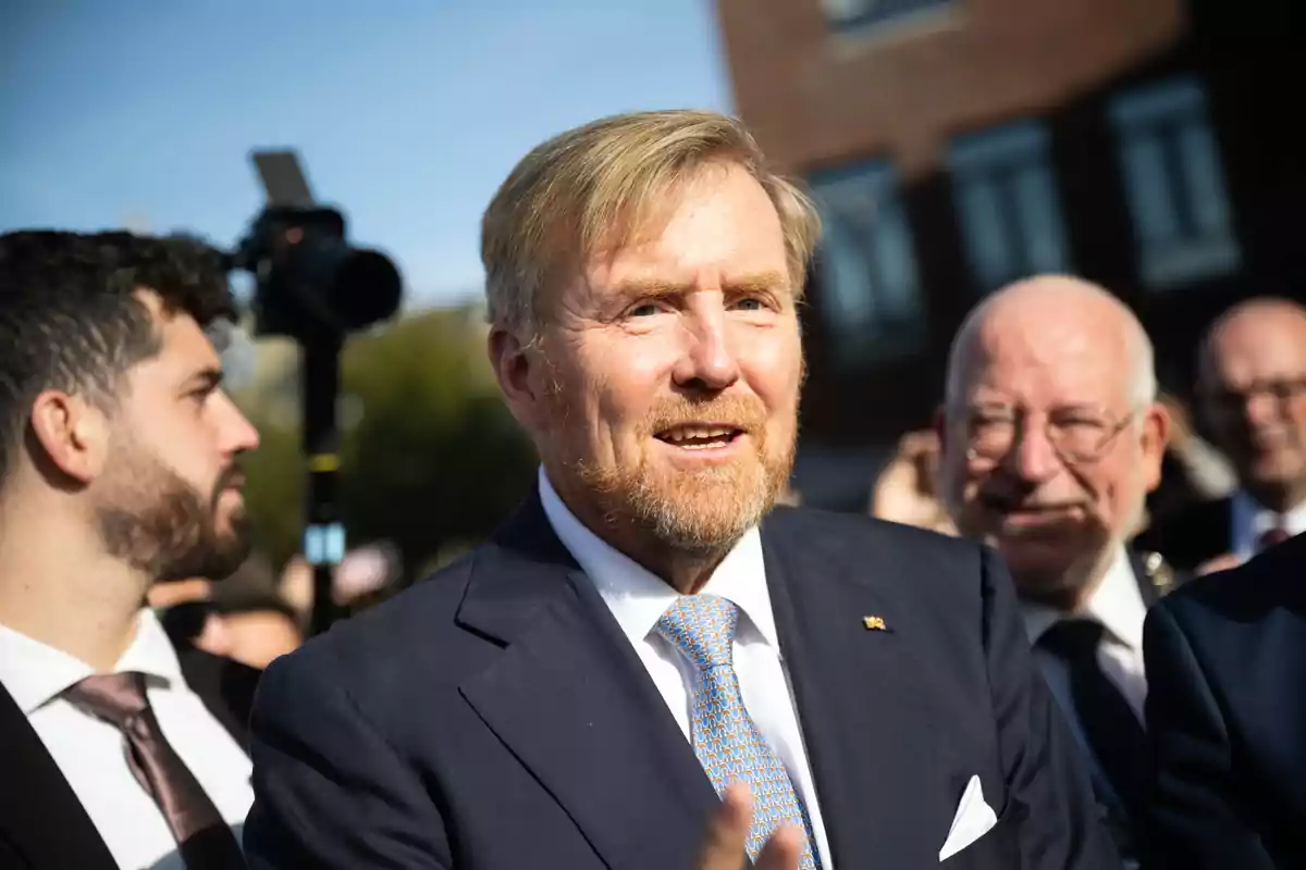 King Willem of the Netherlands, wearing a suit and tie, smiles while surrounded by other people at an outdoor event. King Willem of the Netherlands, wearing a suit and tie, smiles while surrounded by other people at an outdoor event.