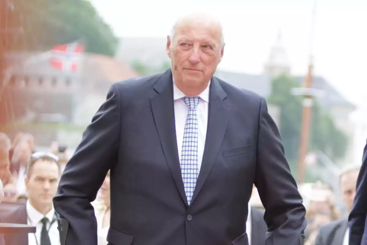 King Harald of Norway in a dark suit and checkered tie, standing outdoors with a Norwegian flag in the background. King Harald of Norway in a dark suit and checkered tie, standing outdoors with a Norwegian flag in the background.