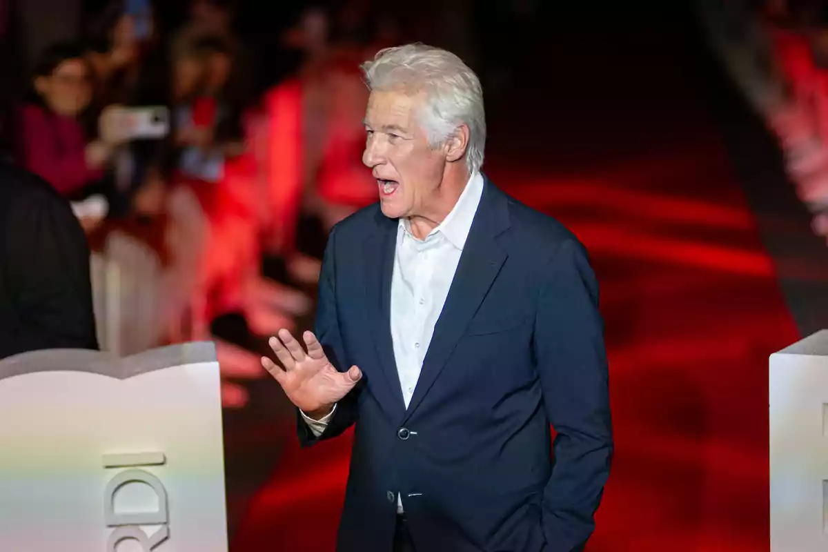 Richard Gere in a dark suit is standing on a red carpet, gesturing with his hand while speaking. Richard Gere in a dark suit is standing on a red carpet, gesturing with his hand while speaking.