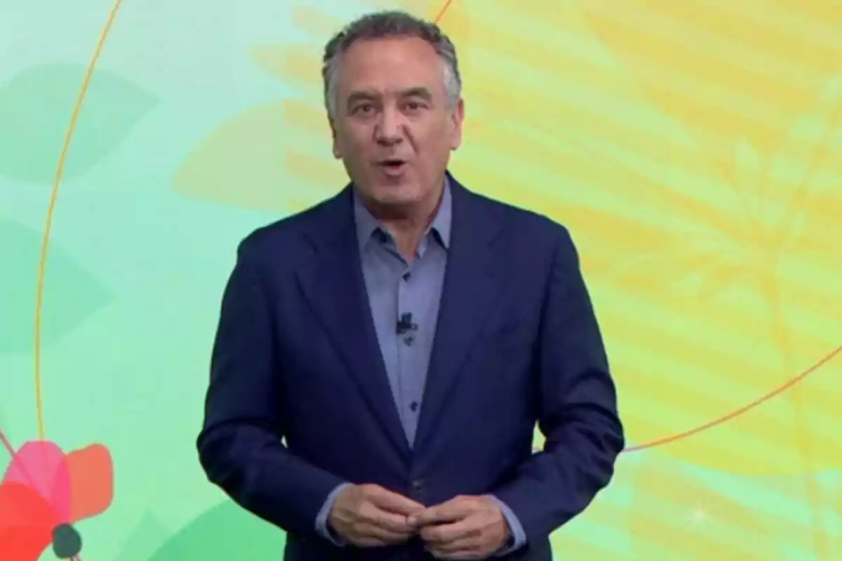 Roberto Brasero vestit amb un vestit blau i camisa grisa dret davant d’un fons acolorit amb formes abstractes i flors, un plató d’Atresmedia. Roberto Brasero vestit amb un vestit blau i camisa grisa dret davant d’un fons acolorit amb formes abstractes i flors, un plató d’Atresmedia.