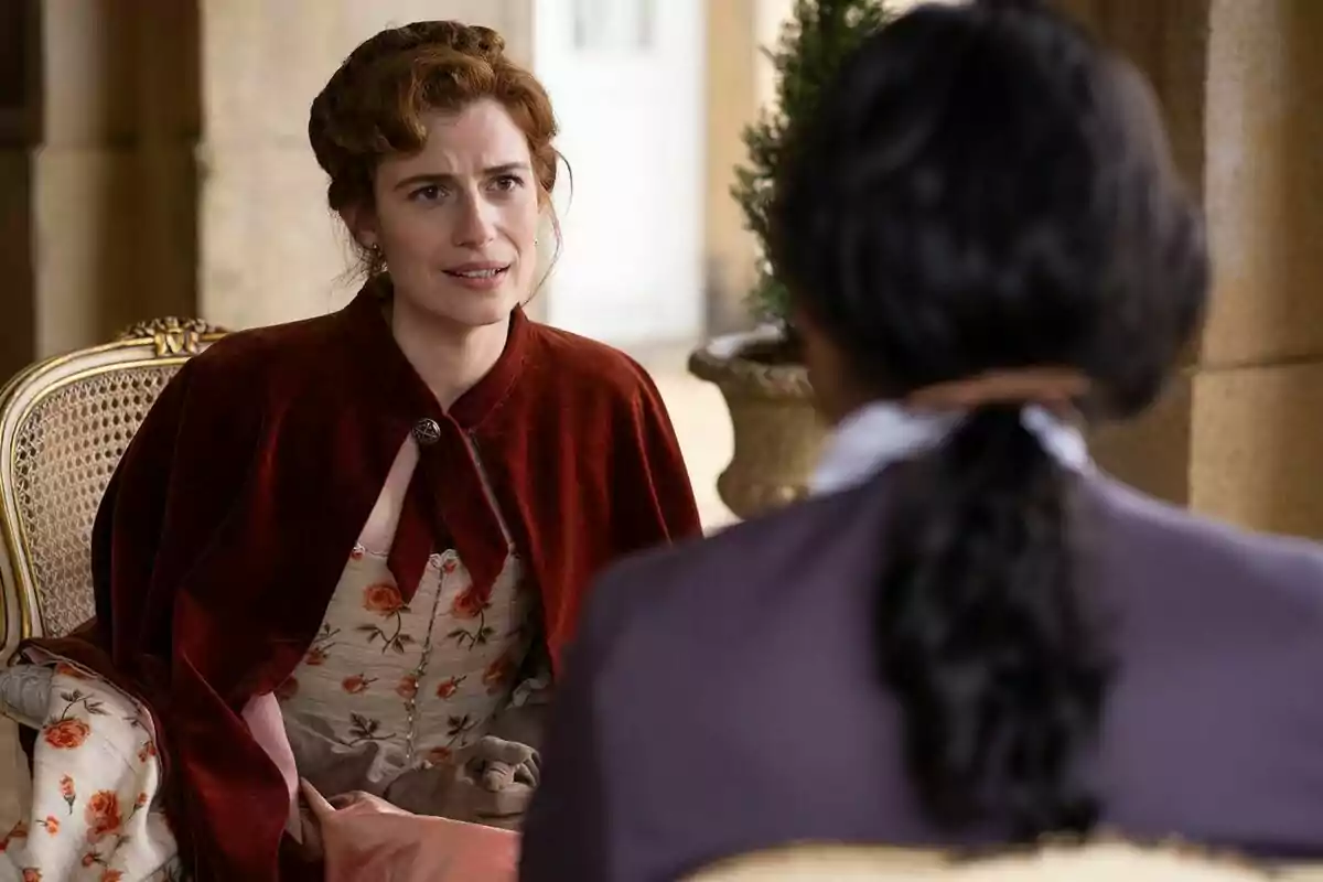 Rocío Suárez de Pugar amb vestit d'època i capa vermella està asseguda en una cadira, conversant amb Marco Pernas d'esquena en un entorn elegant a Valle Salvaje. Rocío Suárez de Pugar amb vestit d'època i capa vermella està asseguda en una cadira, conversant amb Marco Pernas d'esquena en un entorn elegant a Valle Salvaje.