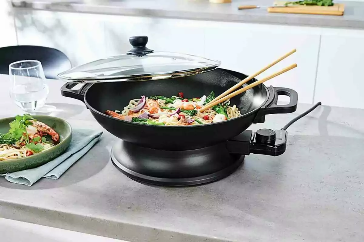Un wok eléctrico con tapa de vidrio cocinando fideos con vegetales y camarones sobre una encimera junto a un plato servido y un vaso de agua. Un wok eléctrico con tapa de vidrio cocinando fideos con vegetales y camarones sobre una encimera junto a un plato servido y un vaso de agua.