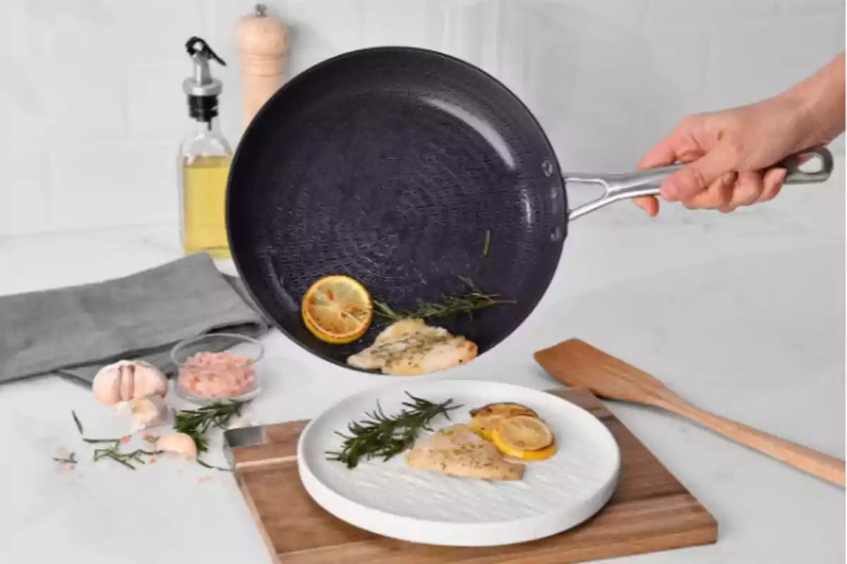 Una paella negra amb mànec de metall aboca filets de peix amb rodanxes de llimona i romaní en un plat blanc sobre una taula de fusta, envoltada d'ingredients com all, sal rosa i oli d'oliva. Una paella negra amb mànec de metall aboca filets de peix amb rodanxes de llimona i romaní en un plat blanc sobre una taula de fusta, envoltada d'ingredients com all, sal rosa i oli d'oliva.