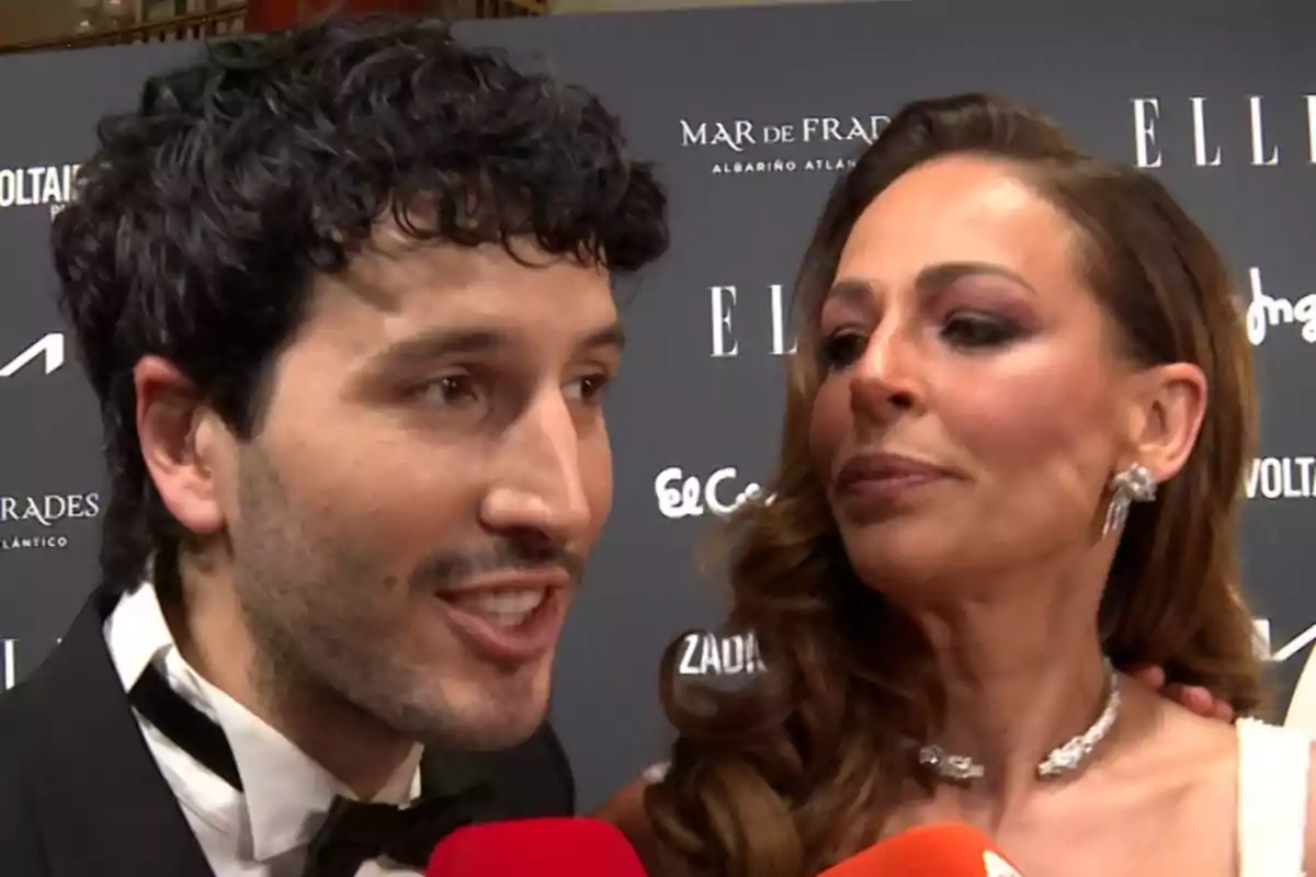 Sebastián Yatra y Eva González elegantemente vestidos posan juntos en una alfombra roja frente a un fondo con logotipos de marcas y micrófonos de prensa. Sebastián Yatra y Eva González elegantemente vestidos posan juntos en una alfombra roja frente a un fondo con logotipos de marcas y micrófonos de prensa.