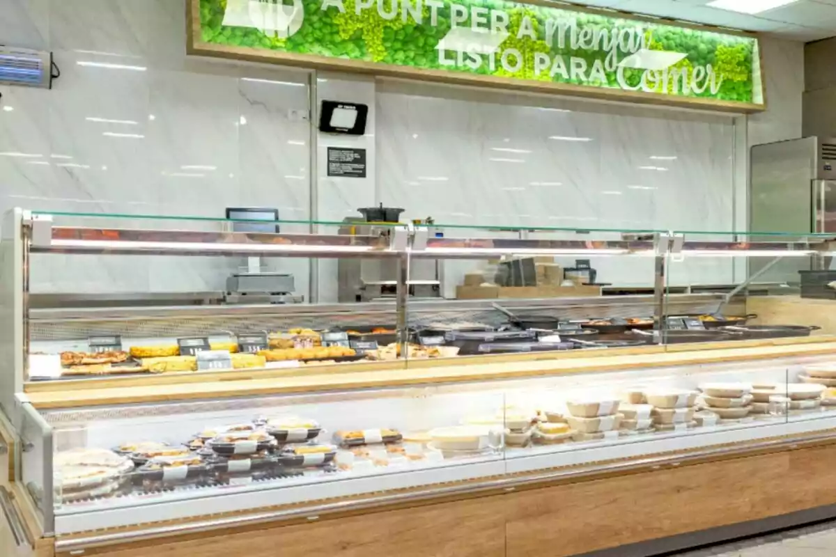 Mostrador de comida preparada en un supermercado con platos y postres exhibidos bajo una vitrina de vidrio.