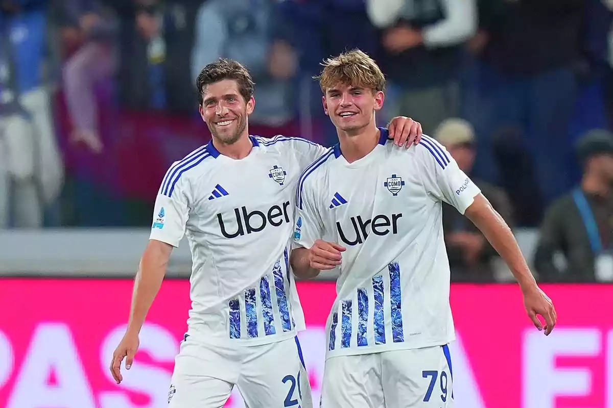 Dos jugadores de fútbol con uniformes blancos y detalles azules sonríen y se abrazan en el campo. Dos jugadores de fútbol con uniformes blancos y detalles azules sonríen y se abrazan en el campo.