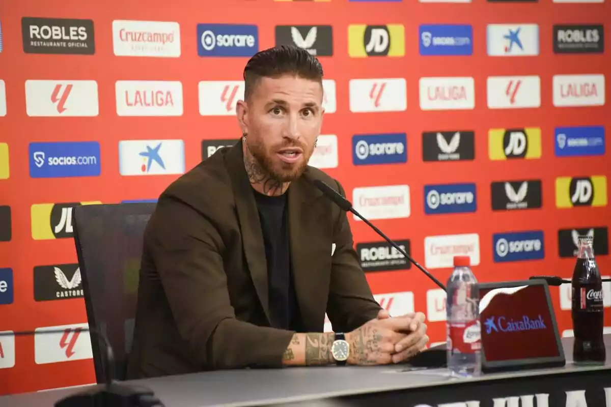 Sergio Ramos con barba y tatuajes en los brazos está hablando en una conferencia de prensa, sentado frente a un micrófono con un fondo lleno de logotipos de patrocinadores. Sergio Ramos con barba y tatuajes en los brazos está hablando en una conferencia de prensa, sentado frente a un micrófono con un fondo lleno de logotipos de patrocinadores.