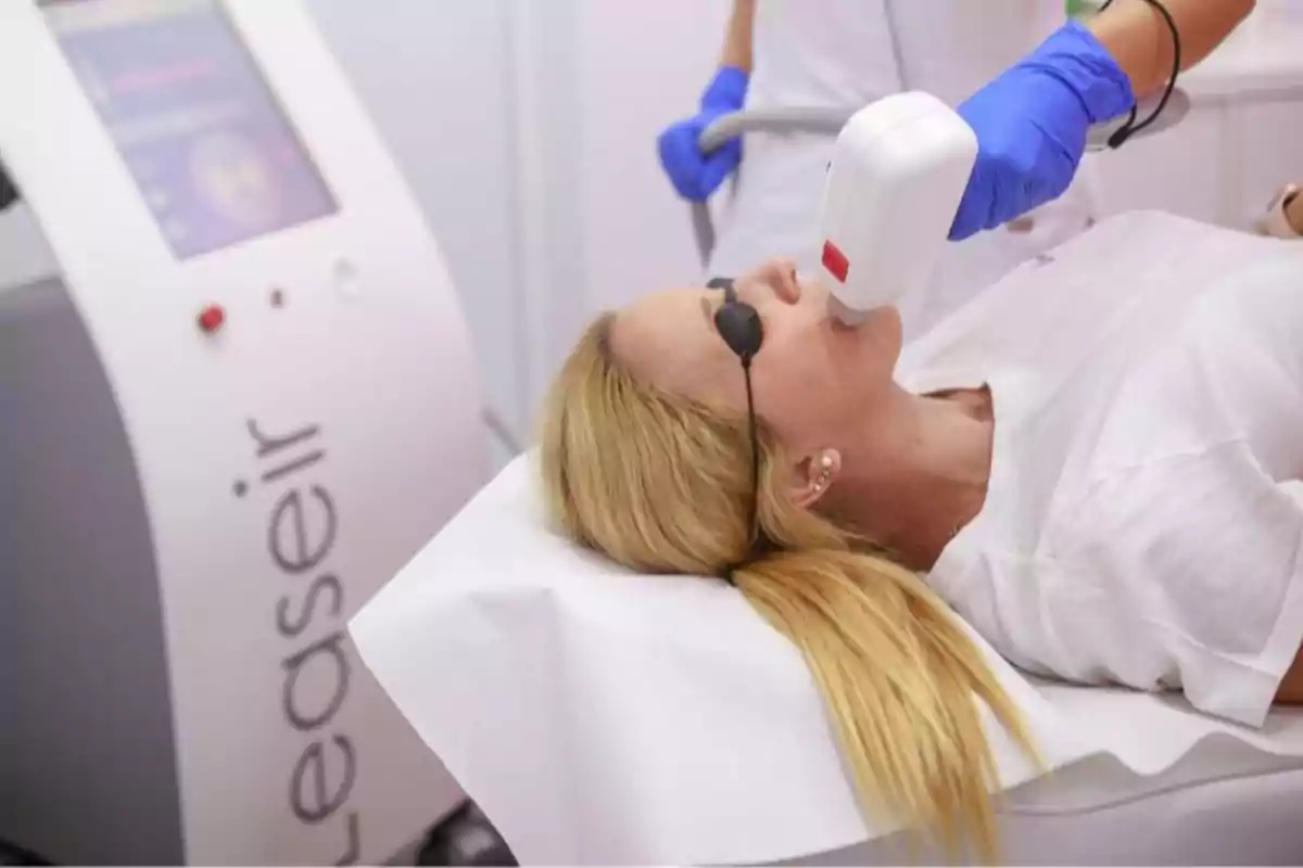 Woman receiving a laser facial treatment at an aesthetic clinic while wearing protective glasses, and a professional with blue gloves holds the device.