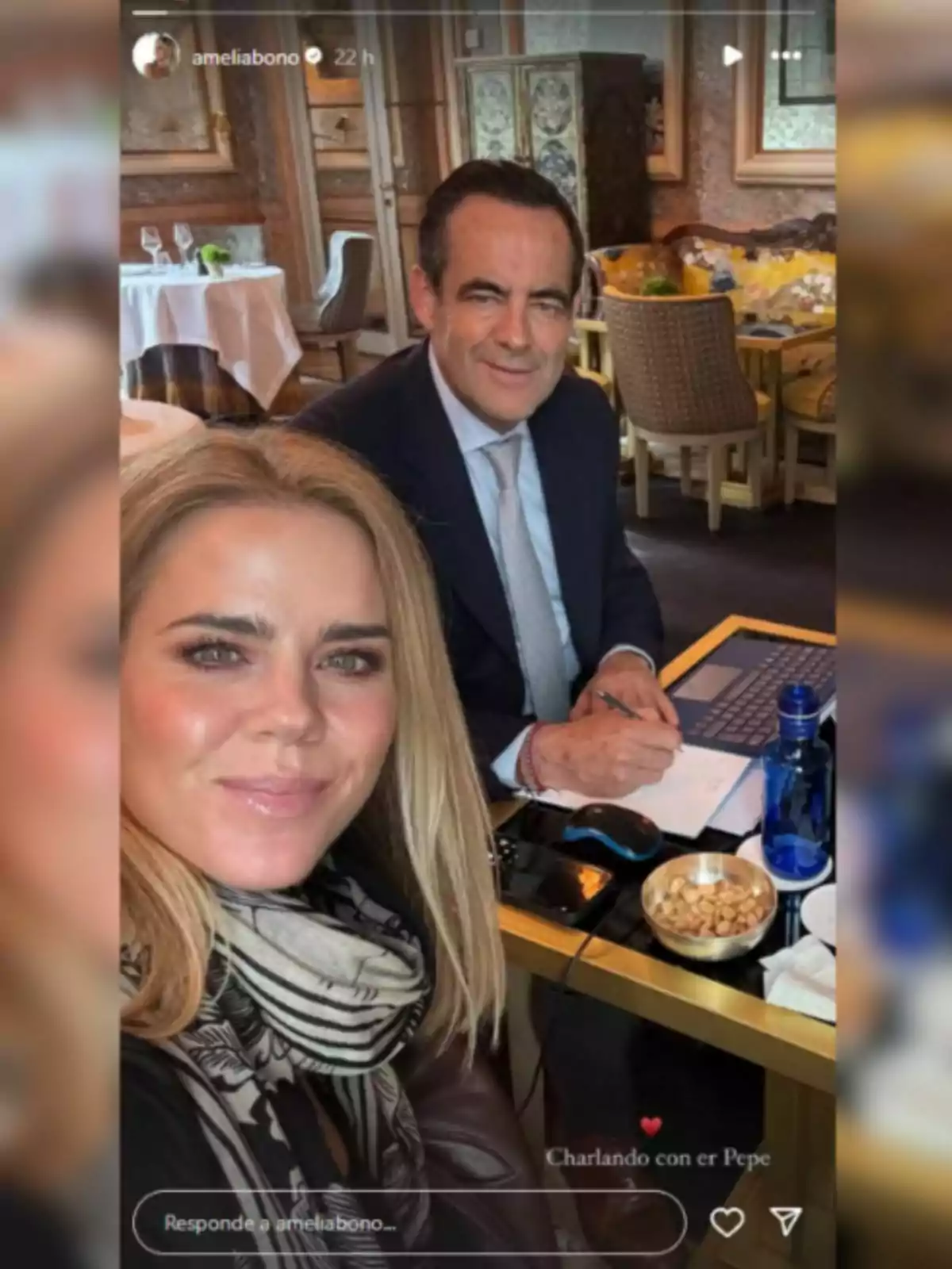 Amelia Bono i José Bono estan asseguts en un restaurant elegant; ella es fa una selfie mentre ell està davant d'un ordinador portàtil amb una llibreta i un bol de fruits secs sobre la taula.