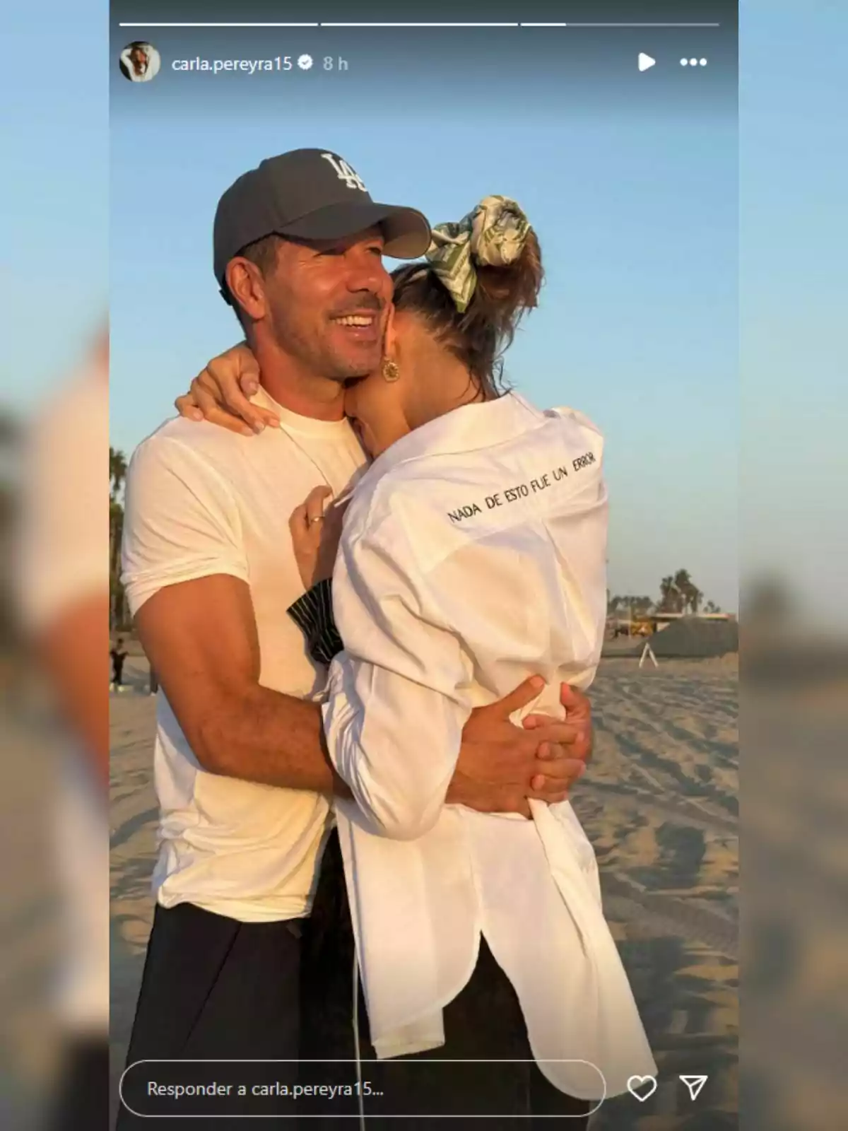 Diego Simeone i Carla Pereyra abraçant-se a la platja al capvespre, tots dos somriuen i la dona porta una camisa blanca amb la frase Diego Simeone i Carla Pereyra abraçant-se a la platja al capvespre, tots dos somriuen i la dona porta una camisa blanca amb la frase