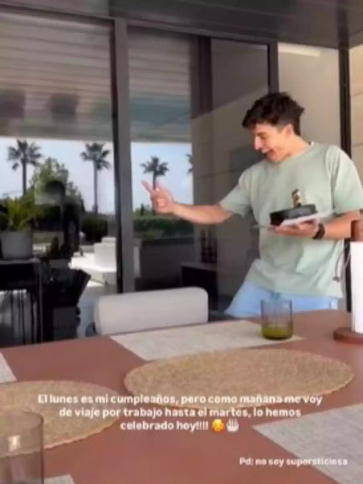 Marc Márquez somrient sosté un pastís petit amb una espelma encesa mentre està dret en una terrassa moderna amb palmeres al fons. A la taula hi ha un got i estovalles individuals. Marc Márquez somrient sosté un pastís petit amb una espelma encesa mentre està dret en una terrassa moderna amb palmeres al fons. A la taula hi ha un got i estovalles individuals.