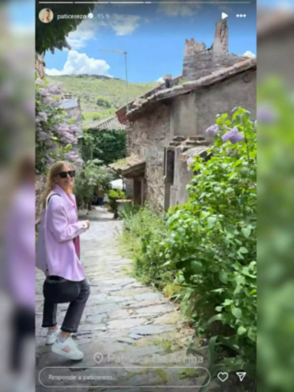 Patricia Cerezo amb ulleres de sol i jaqueta lila camina per un pintoresc camí empedrat envoltat de vegetació i cases de pedra, amb un cel blau i muntanyes al fons. Patricia Cerezo amb ulleres de sol i jaqueta lila camina per un pintoresc camí empedrat envoltat de vegetació i cases de pedra, amb un cel blau i muntanyes al fons.